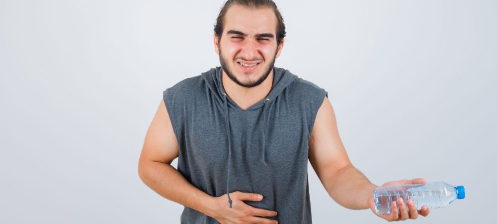 handsome man holding water bottle, having bellyache in sleeveless hoodie, jeans and looking exhausted. front view.