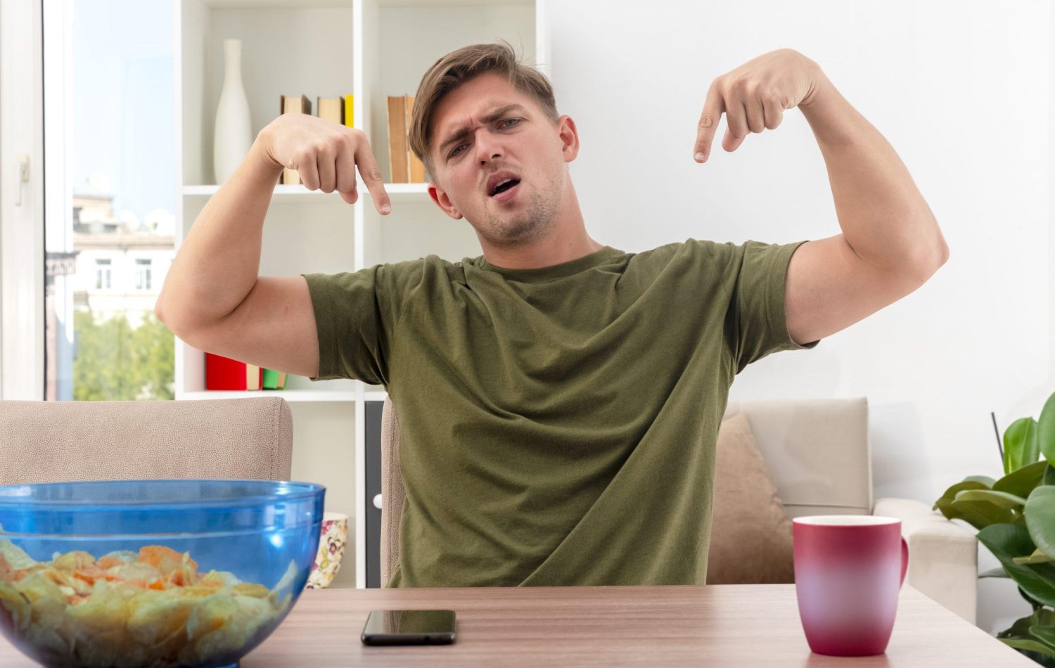 unpleased young blonde handsome man sits at table with bowl of chips phone and cup pointing down with two hands inside design living room